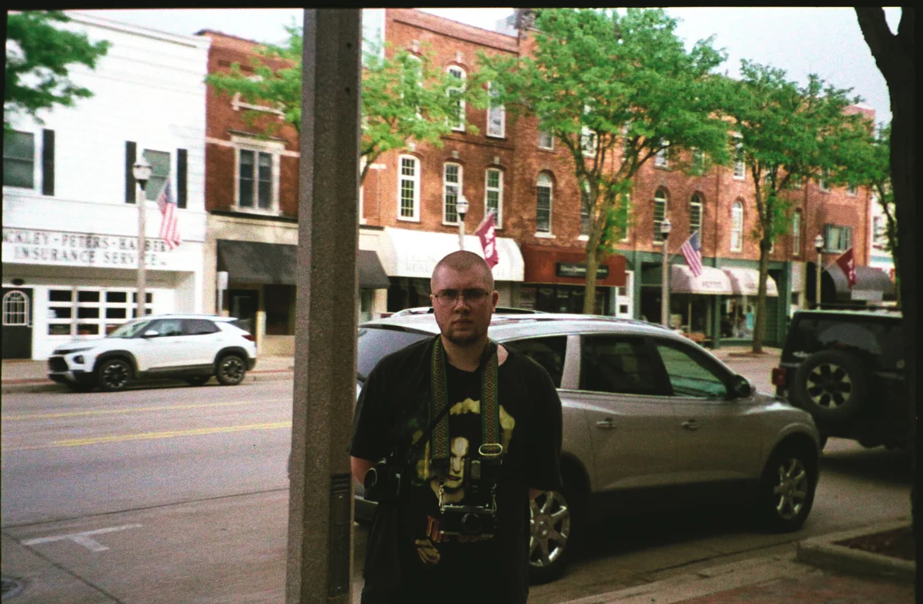 Adrian Harris standing beside a lamppost on a small-town sidewalk, parked cars and brick storefronts behind him.